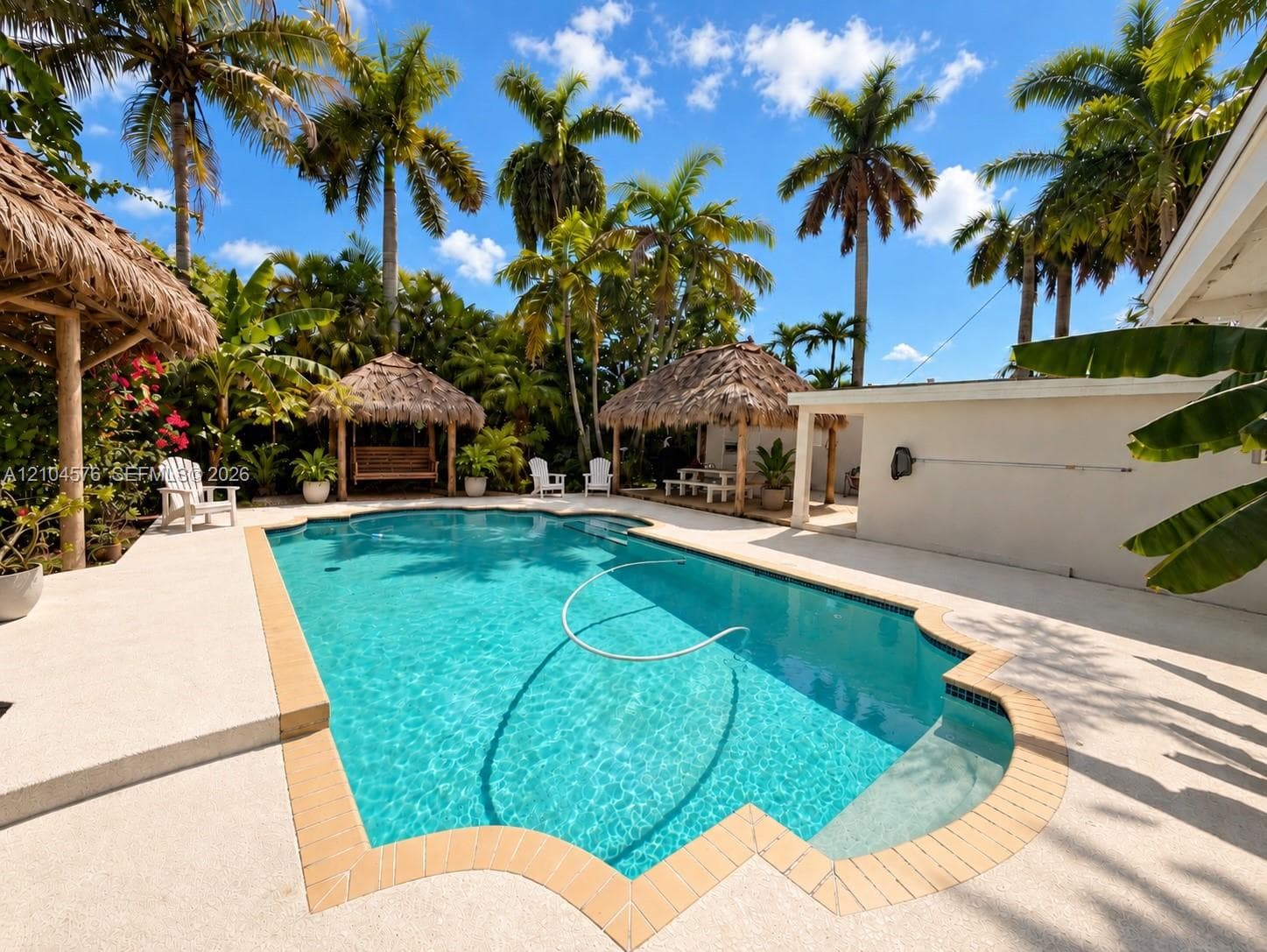 In-ground swimming pool with a tan brick border, surrounded by concrete decking and multiple thatched-roof structures