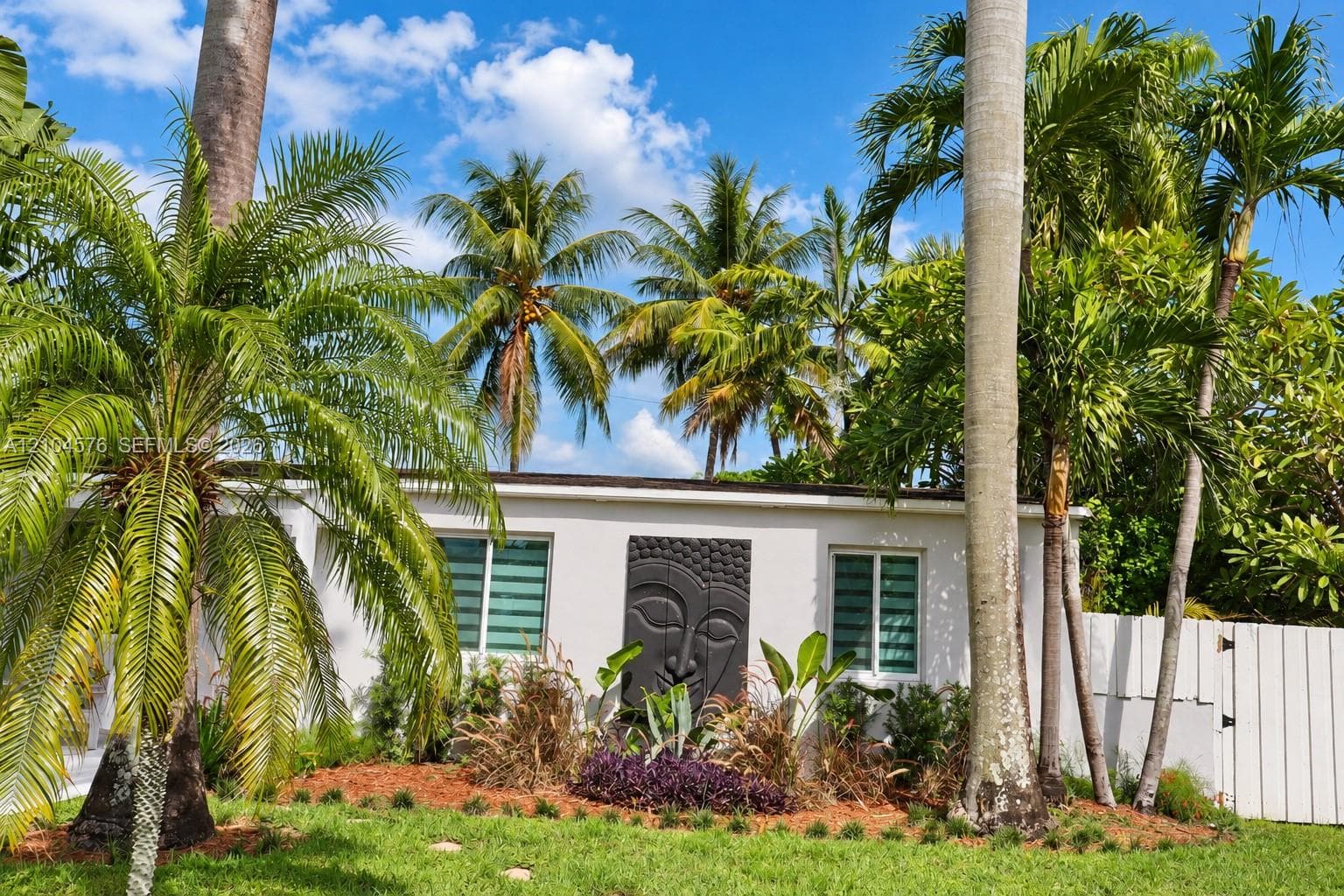 Exterior featuring white stucco facade, manicured landscaping with mature palm trees, decorative wall relief, and perimeter fencing