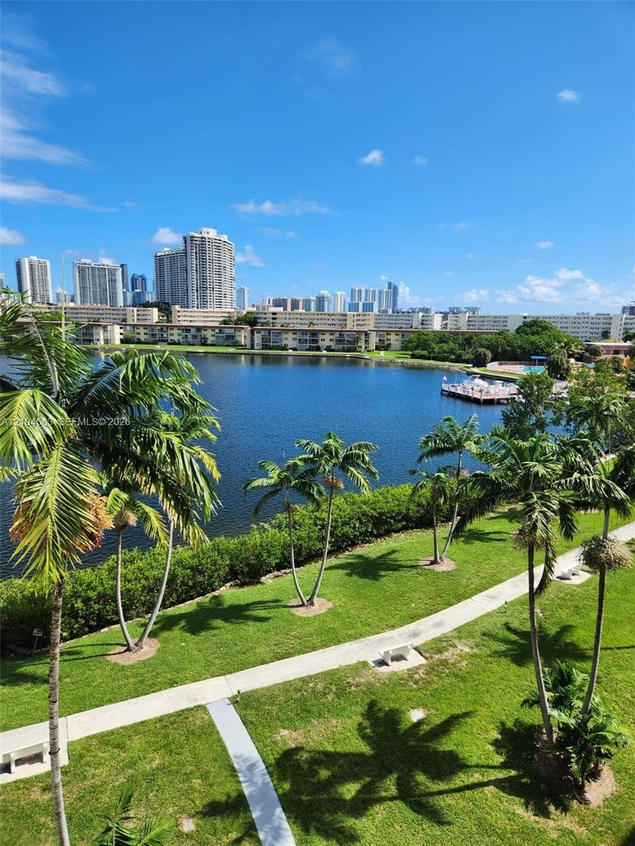Waterfront grounds featuring a concrete walking path, manicured lawn, mature palm trees, and a wooden dock