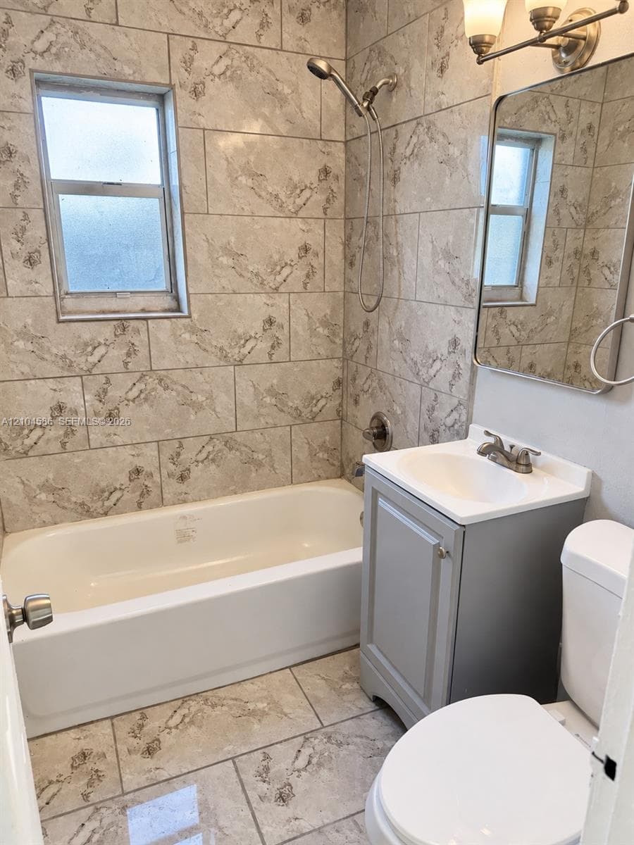 Bathroom featuring full-height tile surround, white porcelain soaking tub, gray-toned vanity with integrated sink, and rectangular wall-mounted mirror