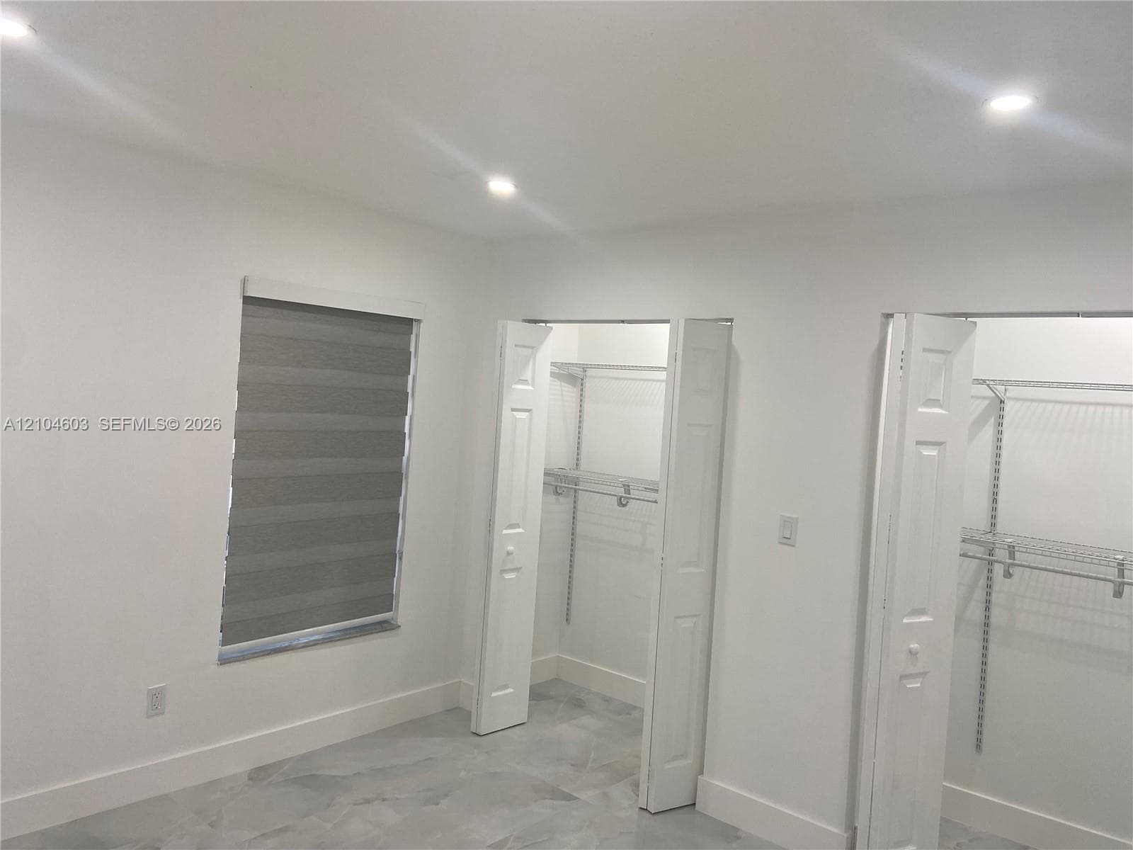 Recessed lighting illuminates the space, featuring a window with layered shades and large bi-fold closet doors
