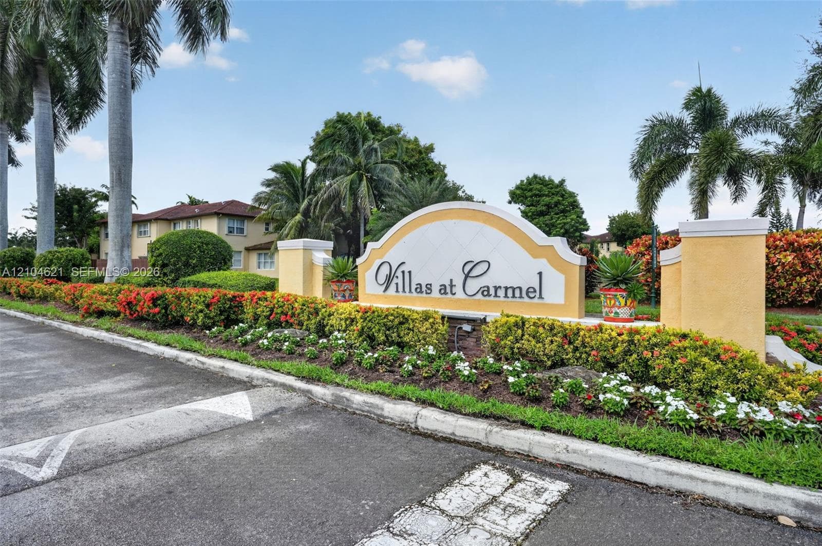 Entrance signage featuring stucco monuments, landscaped flower beds, mature palm trees, and paved asphalt road access