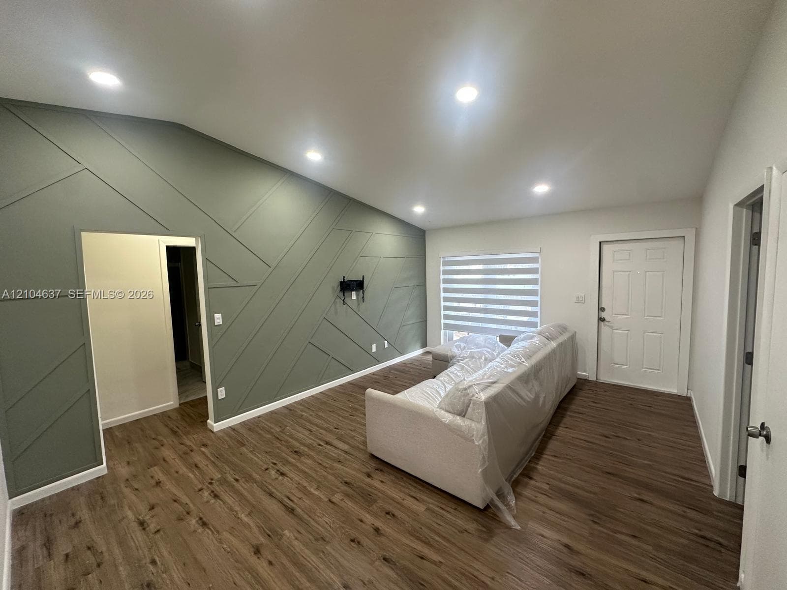 Vaulted ceiling with recessed lighting, accent wall with geometric paneling, wood-finish flooring, and a window with horizontal blinds