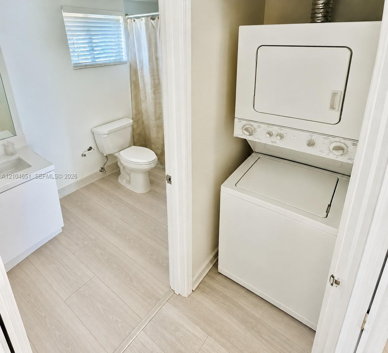 Integrated laundry closet featuring a stacked washer and dryer, wood-finish flooring, white vanity with an integrated basin, and a bathroom equipped with a porcelain toilet and shower combination
