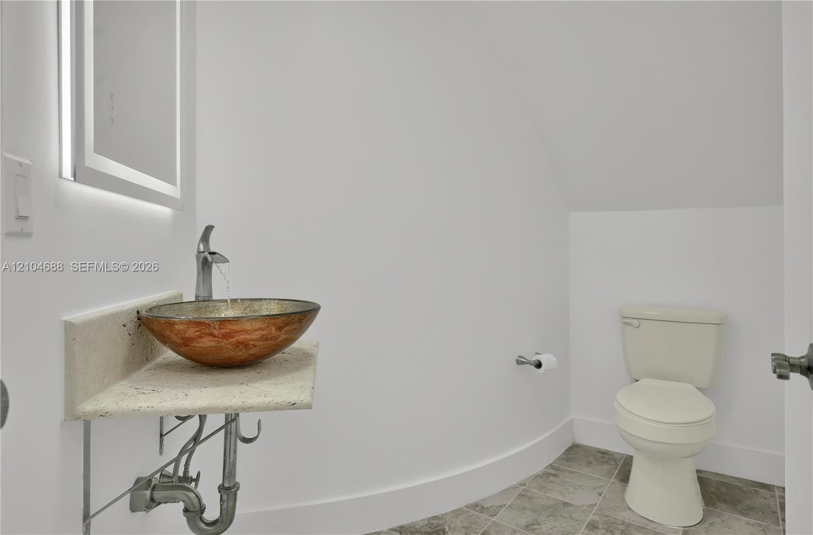 Wall-mounted floating stone vanity featuring a circular copper-tone vessel sink, brushed metal faucet, integrated LED-backlit mirror, and grey tile flooring