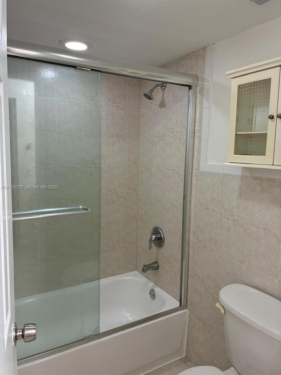 Bathtub and shower combination featuring a sliding glass enclosure, neutral tile surround, recessed lighting, and built-in upper wall cabinetry