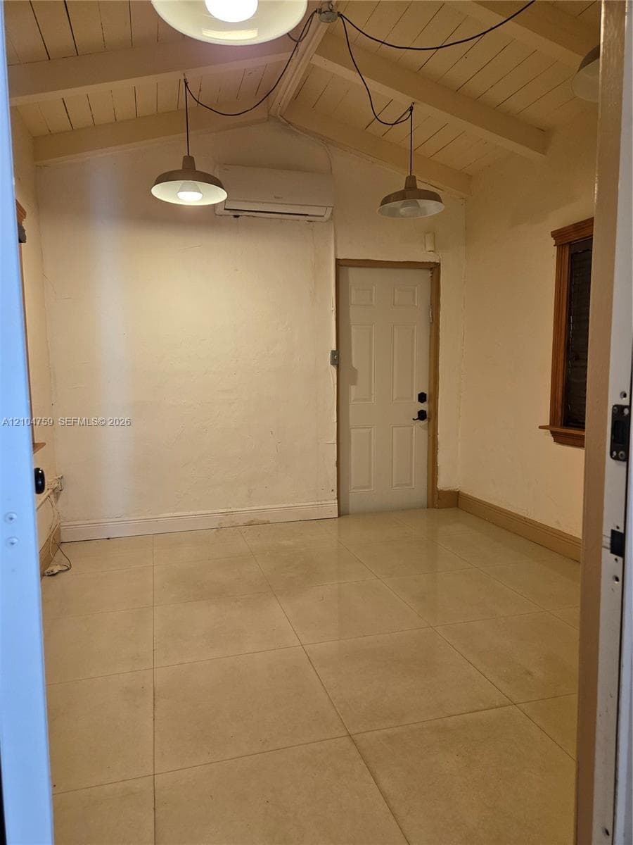 Room with neutral tile flooring, exposed vaulted beam ceiling, wall-mounted ductless air conditioning unit, and dual industrial-style pendant lighting