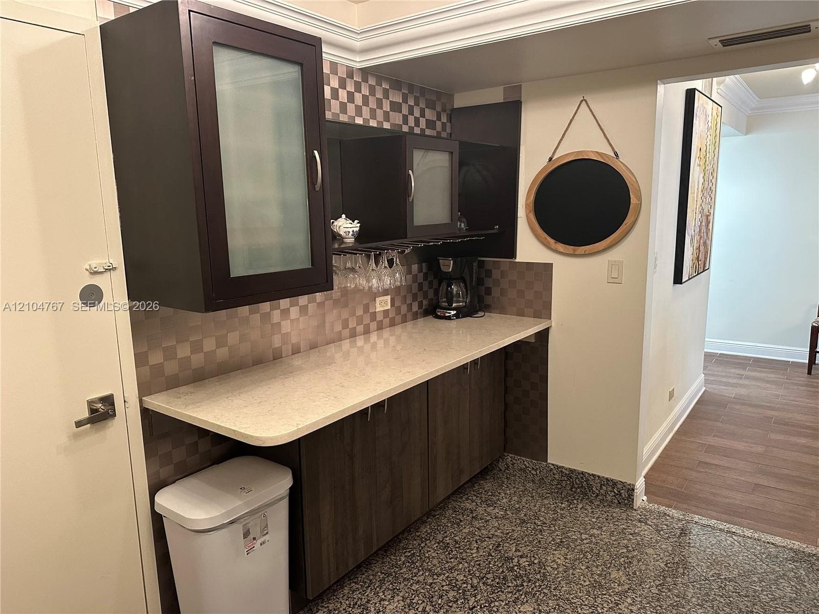 Built-in cabinetry featuring dark wood-finish lower cabinets, upper cabinets with frosted glass inserts, a light-toned solid surface countertop, and a metallic checkered backsplash