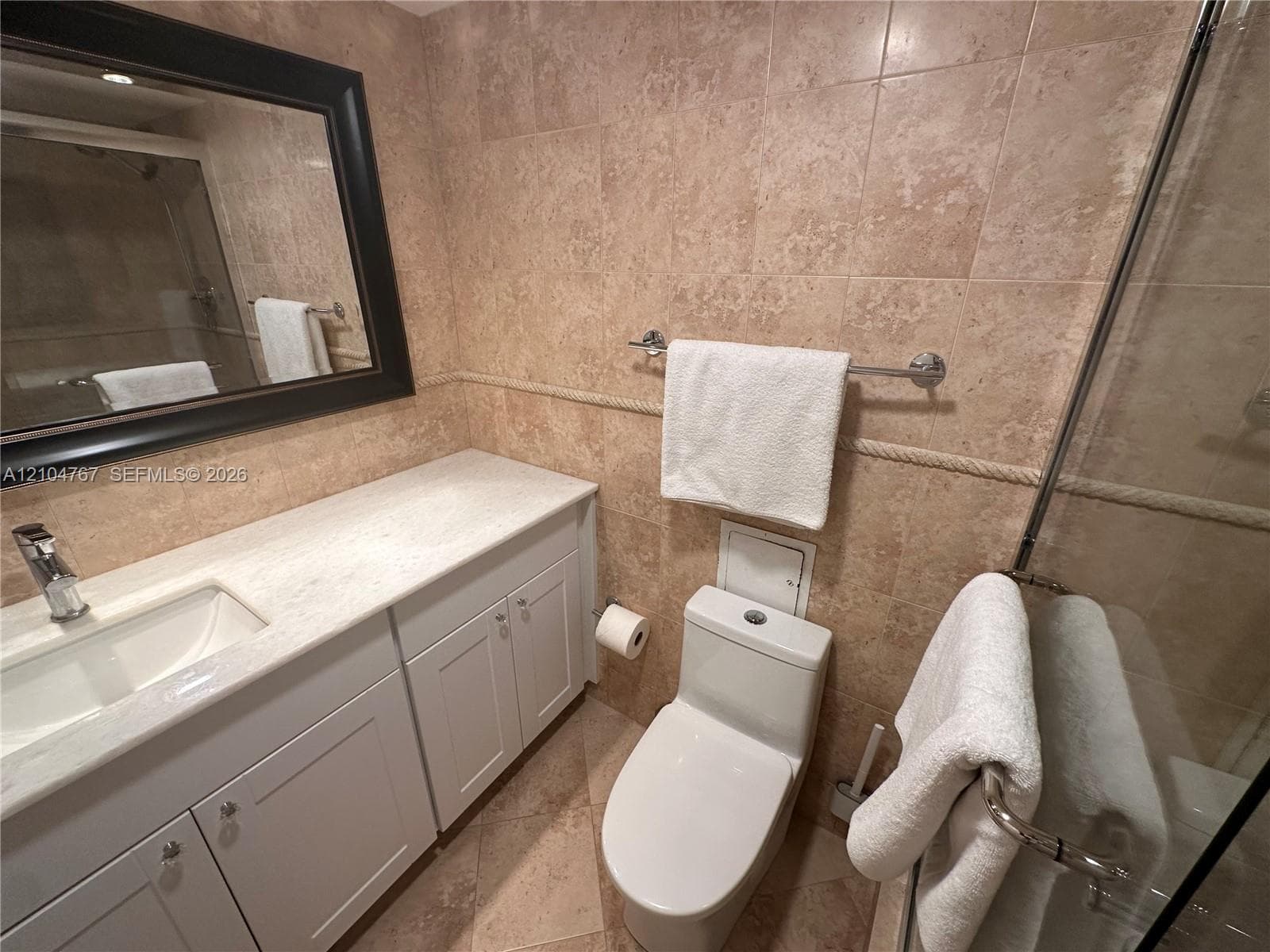 Bathroom featuring a white vanity cabinet with a light-toned countertop, undermount sink, ceramic wall tiling with rope-style decorative trim, and a glass-enclosed shower