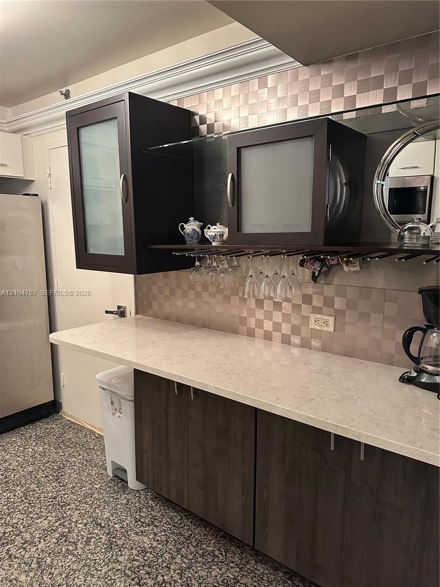 Dark wood-finish cabinetry with frosted glass inserts, stone-finish countertops, metallic checkered backsplash, integrated stemware rack, and crown molding