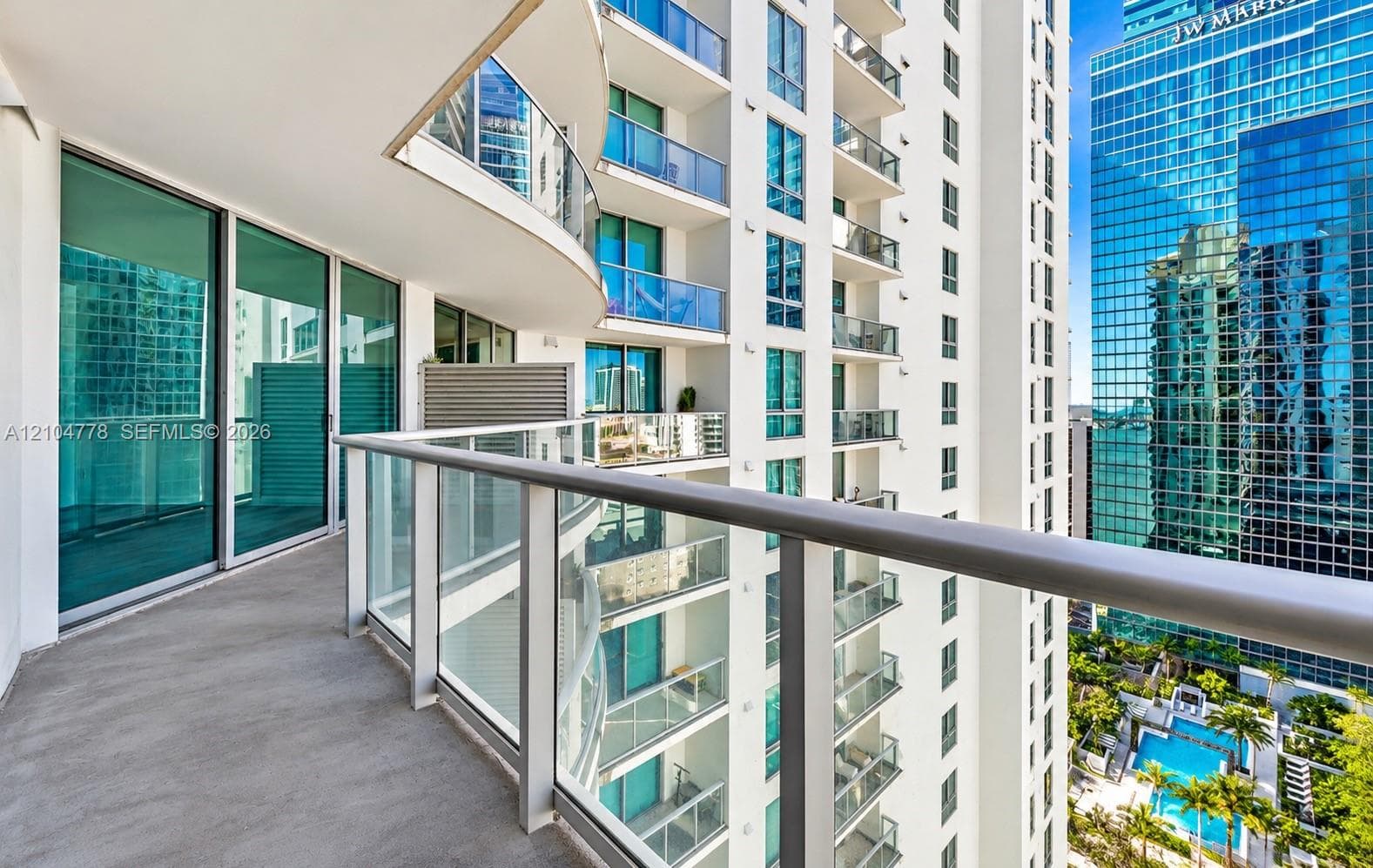 Curved balcony with glass railing, sliding glass doors, concrete flooring, and surrounding high-rise architecture