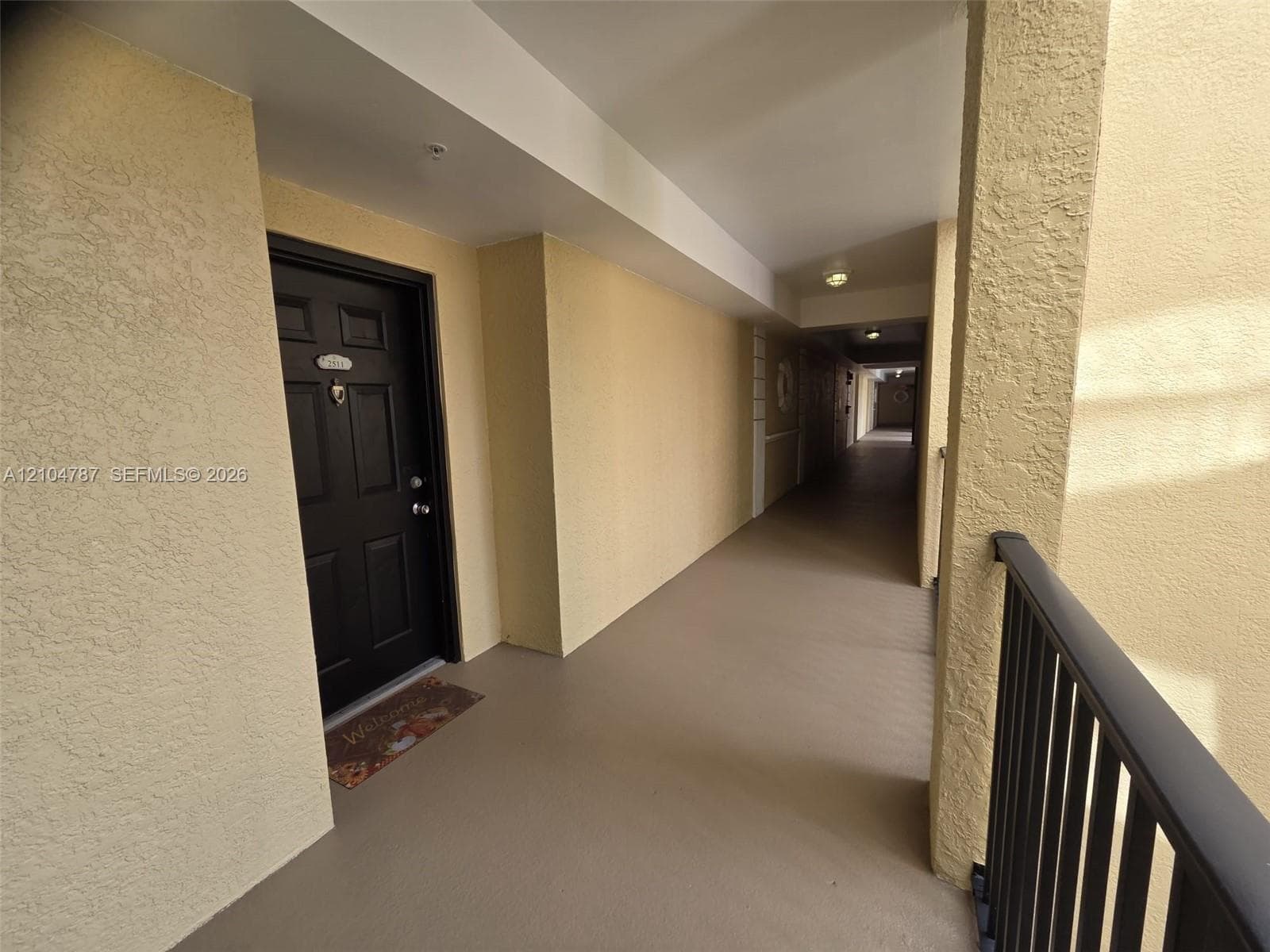 Covered exterior walkway featuring textured stucco walls, black paneled entry door with hardware, flush-mount ceiling lighting, and black metal railings