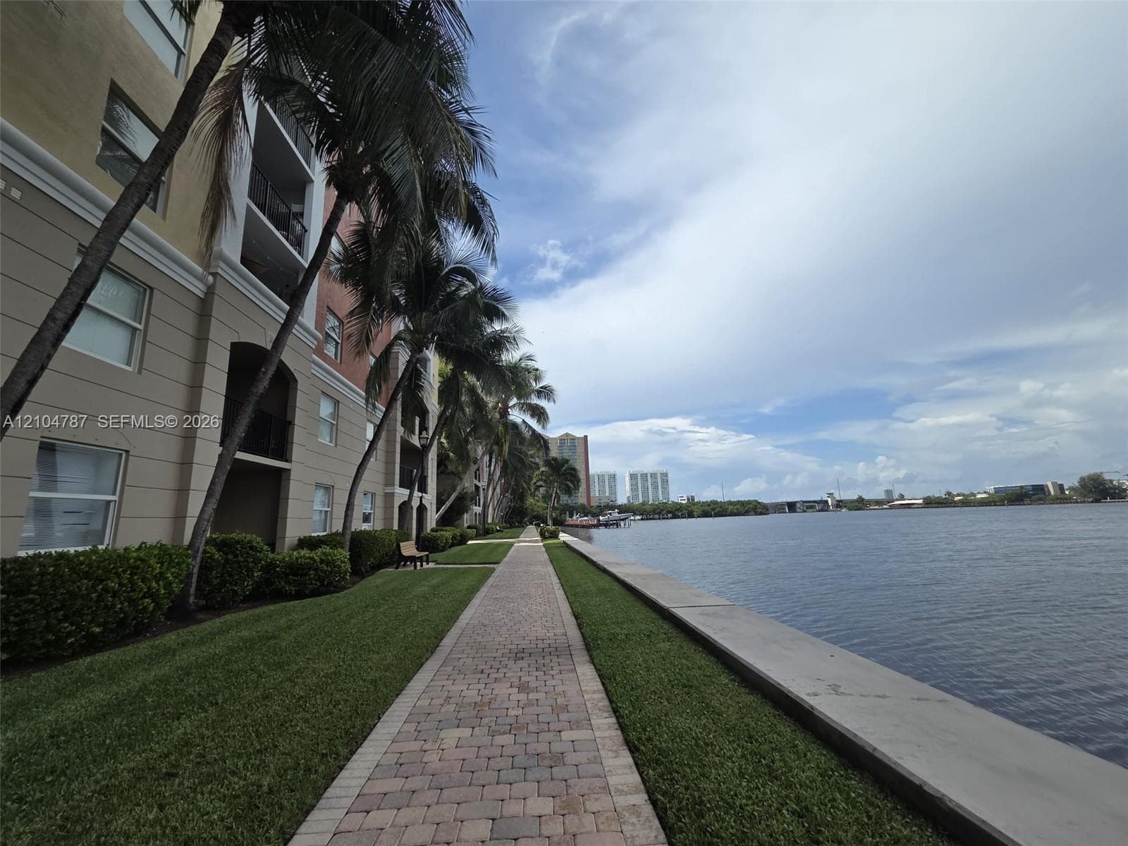 Waterfront walkway featuring paver-style hardscaping, manicured grass borders, and mature palm trees
