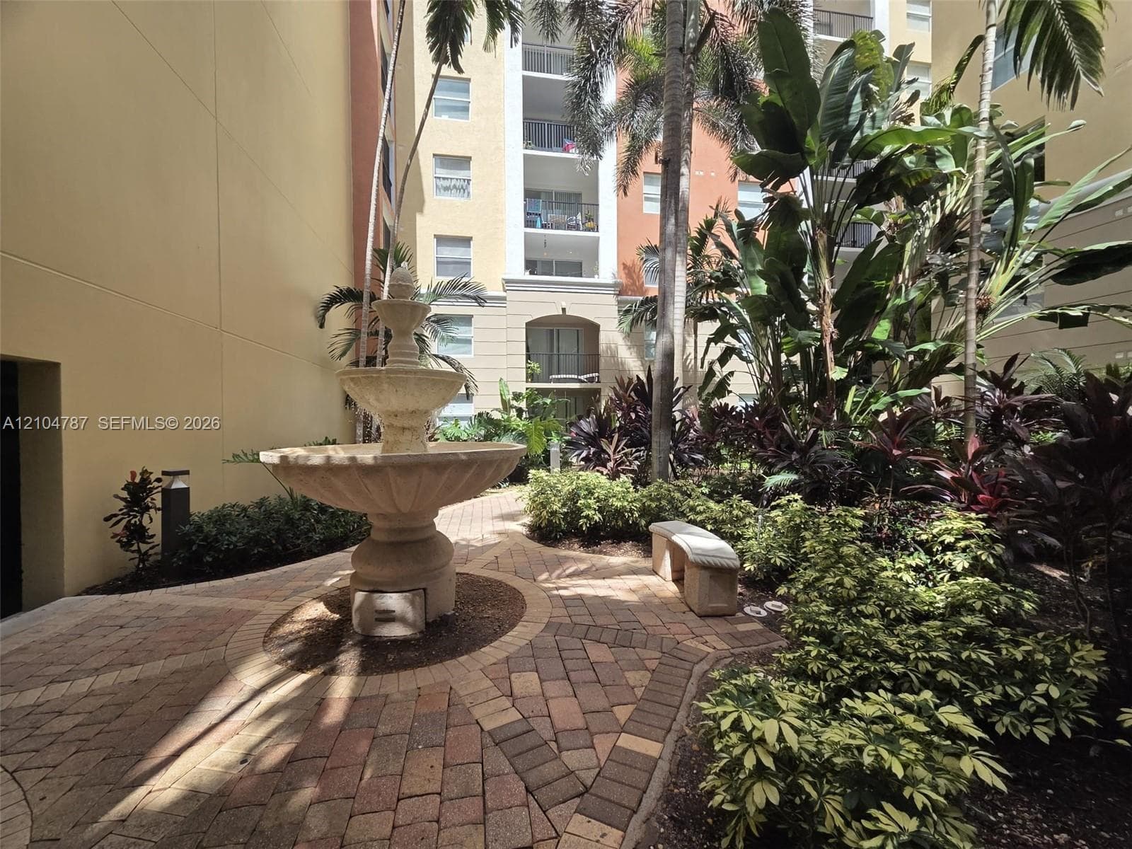 Courtyard featuring a tiered stone fountain, decorative paver hardscaping, mature tropical landscaping, and a permanent stone bench