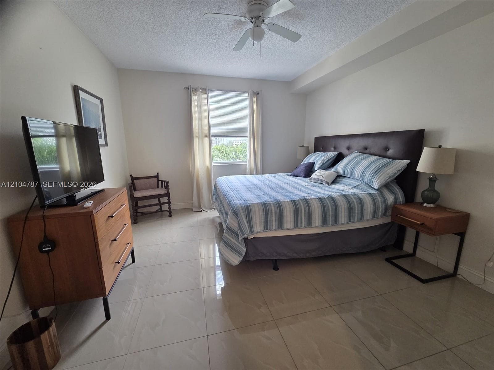 Neutral-toned room featuring large-format ceramic tile flooring, a centered ceiling fan with integrated lighting, and a single window with adjustable blinds