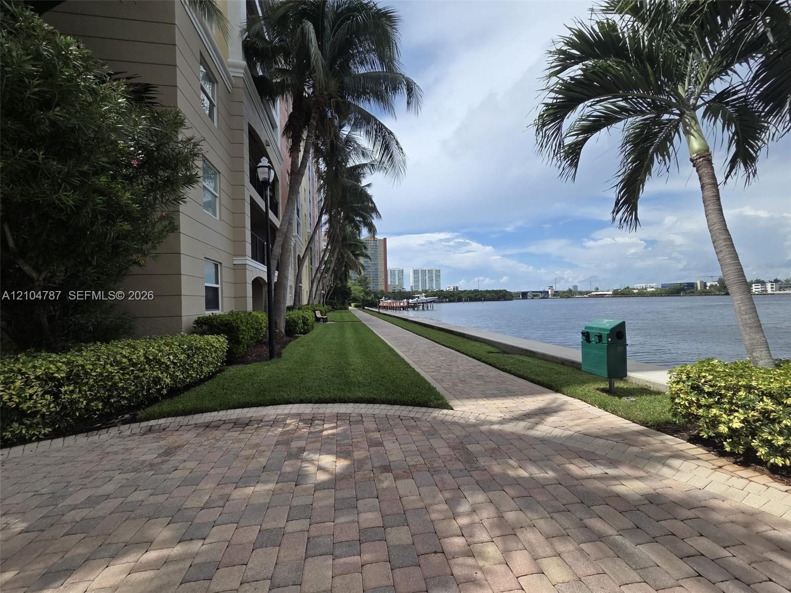 Waterfront grounds featuring a paver walkway, manicured lawn, established palm trees, and a concrete bulkhead