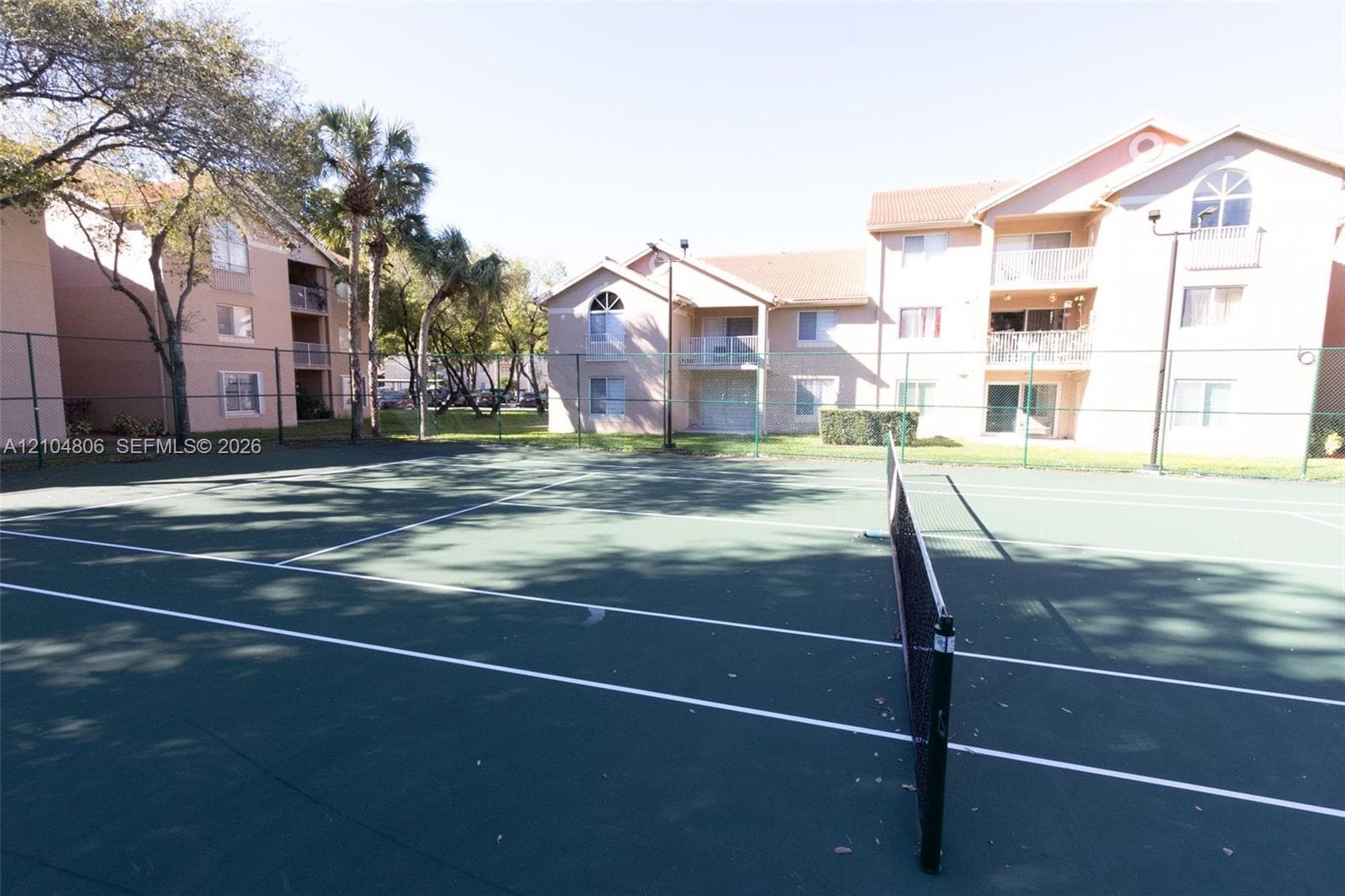 Hardcourt tennis court with net, surrounded by fencing and mature trees