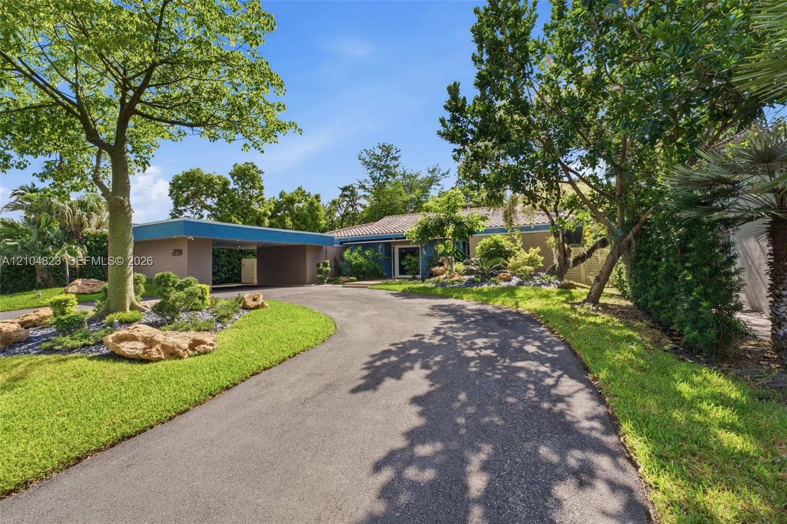 Paved circular driveway leading to a covered carport, mature trees, manicured lawn, and established landscaping