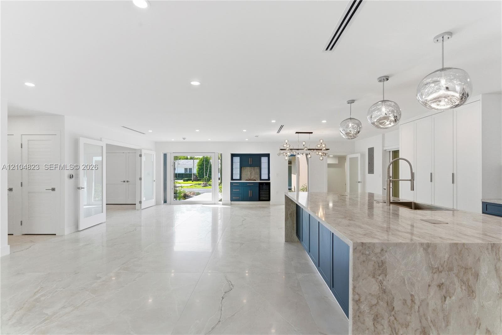 Open-concept kitchen featuring an expansive waterfall-edge island, stone-finish countertops, custom cabinetry, recessed lighting, and glass pendant fixtures