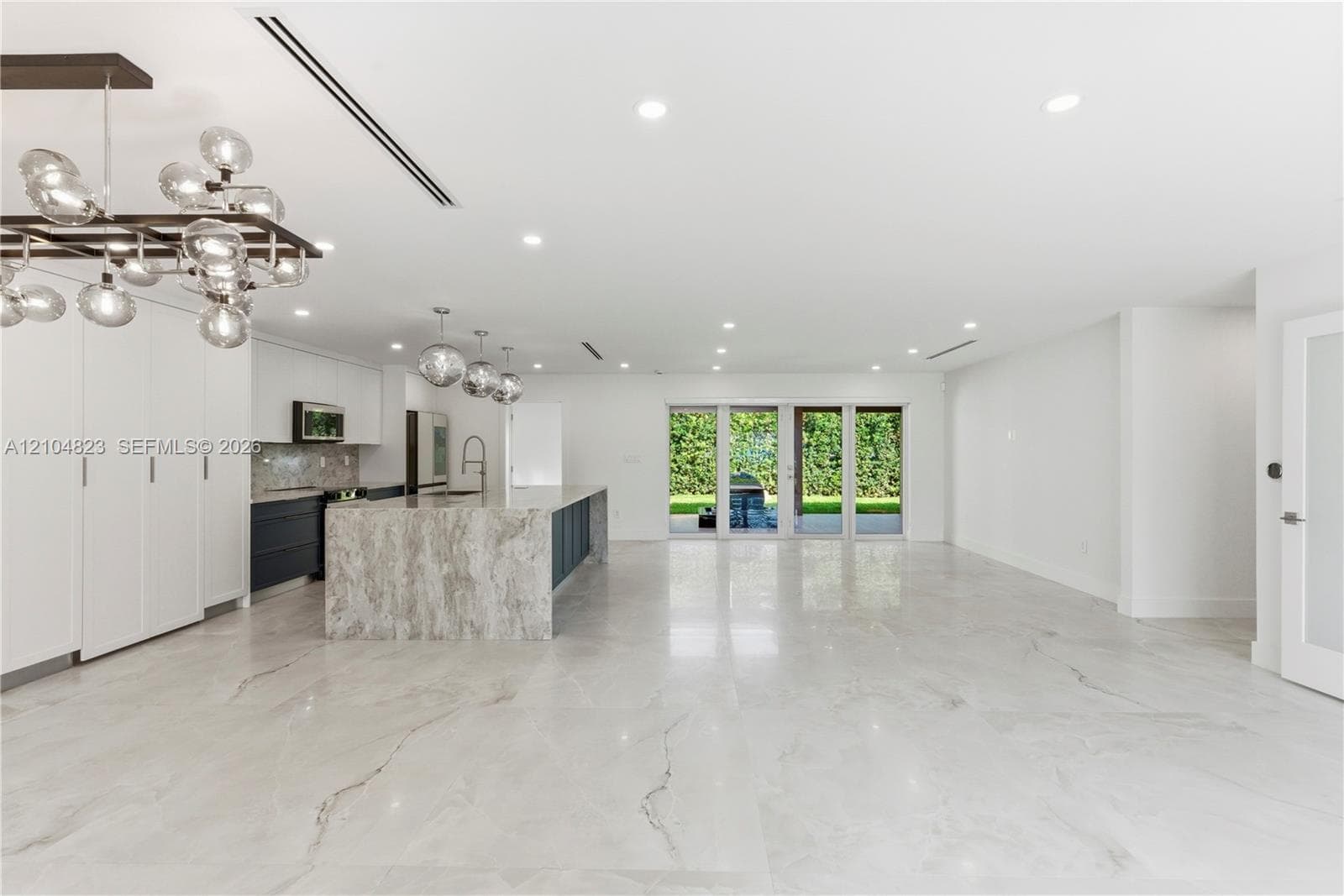 Open-concept interior featuring large-format tile flooring, a stone-surround waterfall edge island, custom cabinetry, contemporary multi-globe light fixtures, and glass patio doors