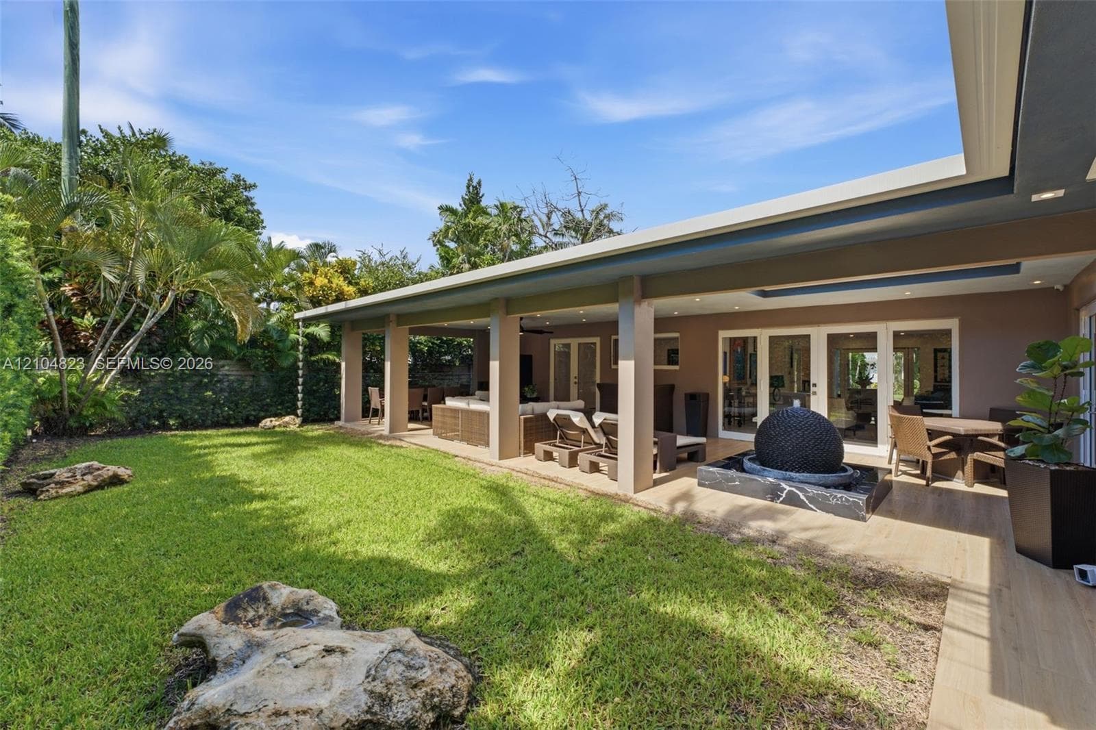 Expansive green lawn with mature landscaping, covered patio with recessed lighting, and a modern water feature