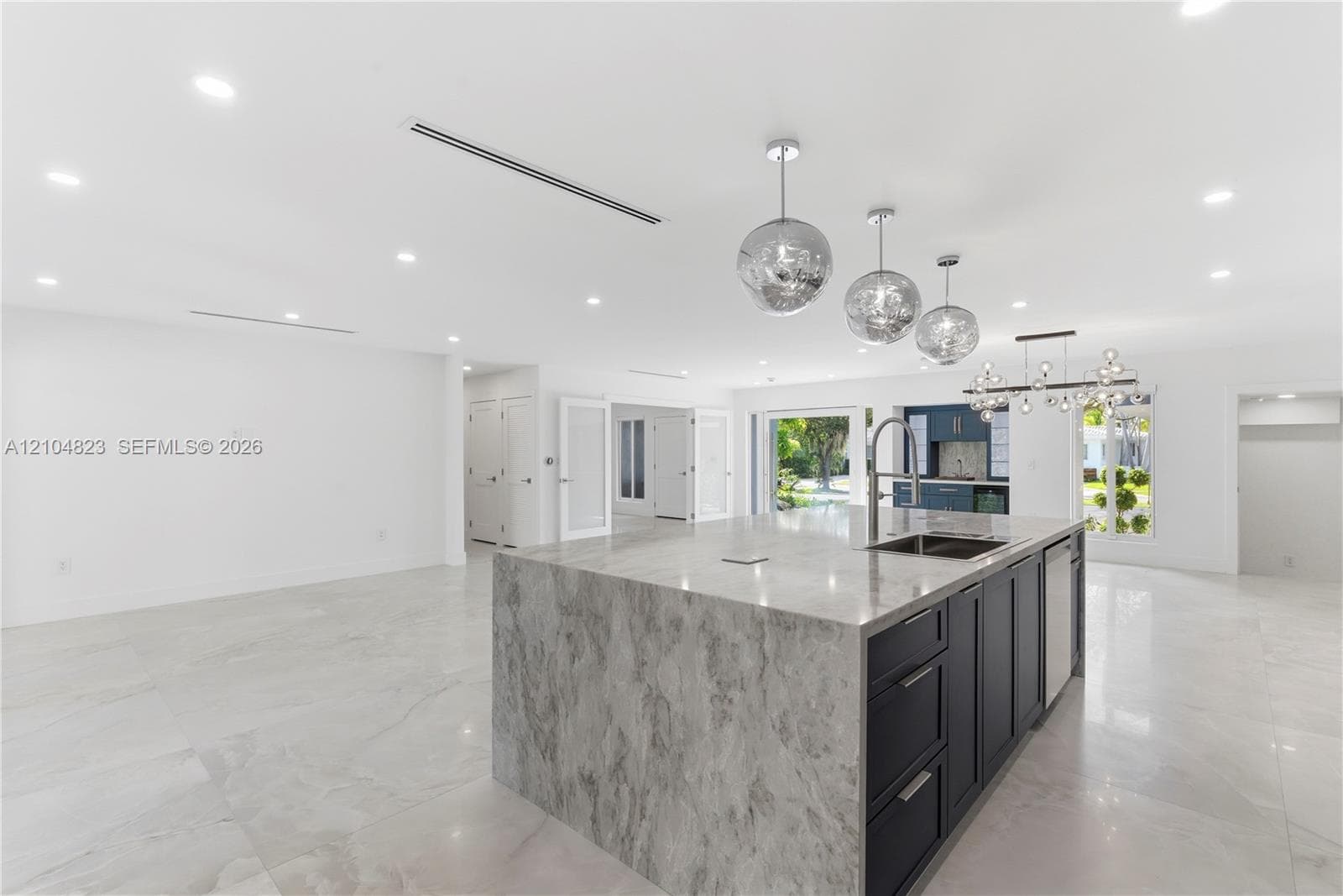 Kitchen featuring a waterfall-edge island with stone-finish countertop, dark-toned cabinetry, recessed lighting, metallic pendant fixtures, and large-format tile flooring