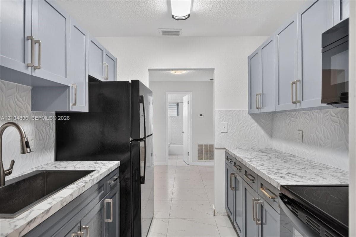 Kitchen featuring light blue shaker-style cabinetry, stone-patterned countertops, textured tile backsplash, dark-finished hardware, and ceramic tile flooring