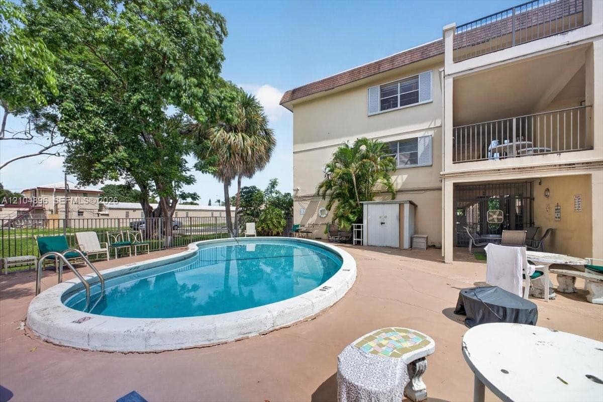 Kidney-shaped in-ground swimming pool with metal ladder, expansive concrete pool deck, multi-level exterior balconies with metal railings, and mature palm trees