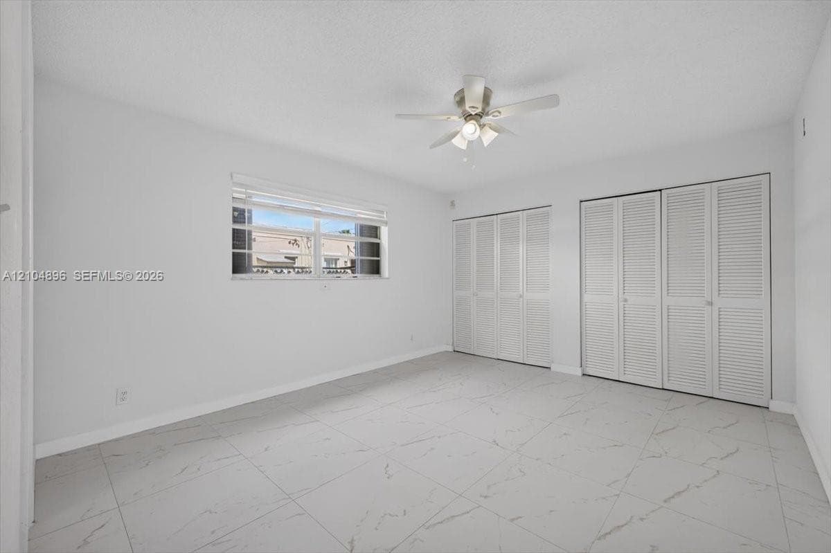 Large-format tile flooring with marble-style veining, dual sets of louvered-door closets, and a ceiling-mounted light fixture with fan