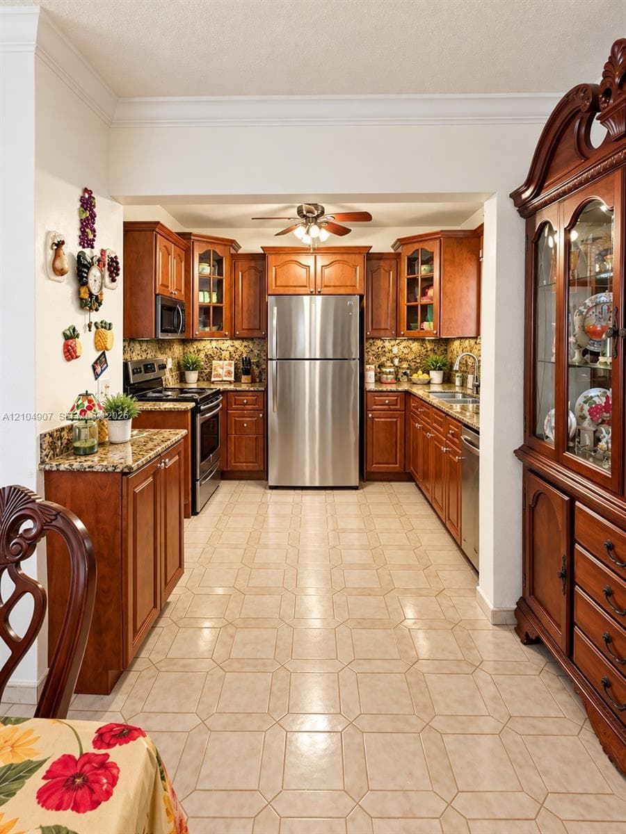 Kitchen featuring medium-toned wood cabinetry, stainless steel appliances, stone-patterned countertops, tile flooring, and under-cabinet lighting