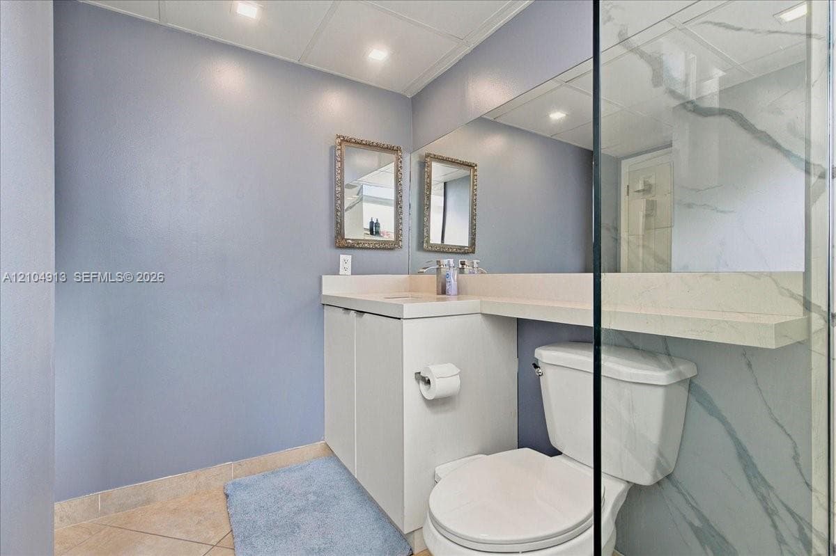 Built-in vanity featuring a light-toned countertop, large wall-mounted mirror, glass-enclosed shower with stone-finish surround, and tile flooring