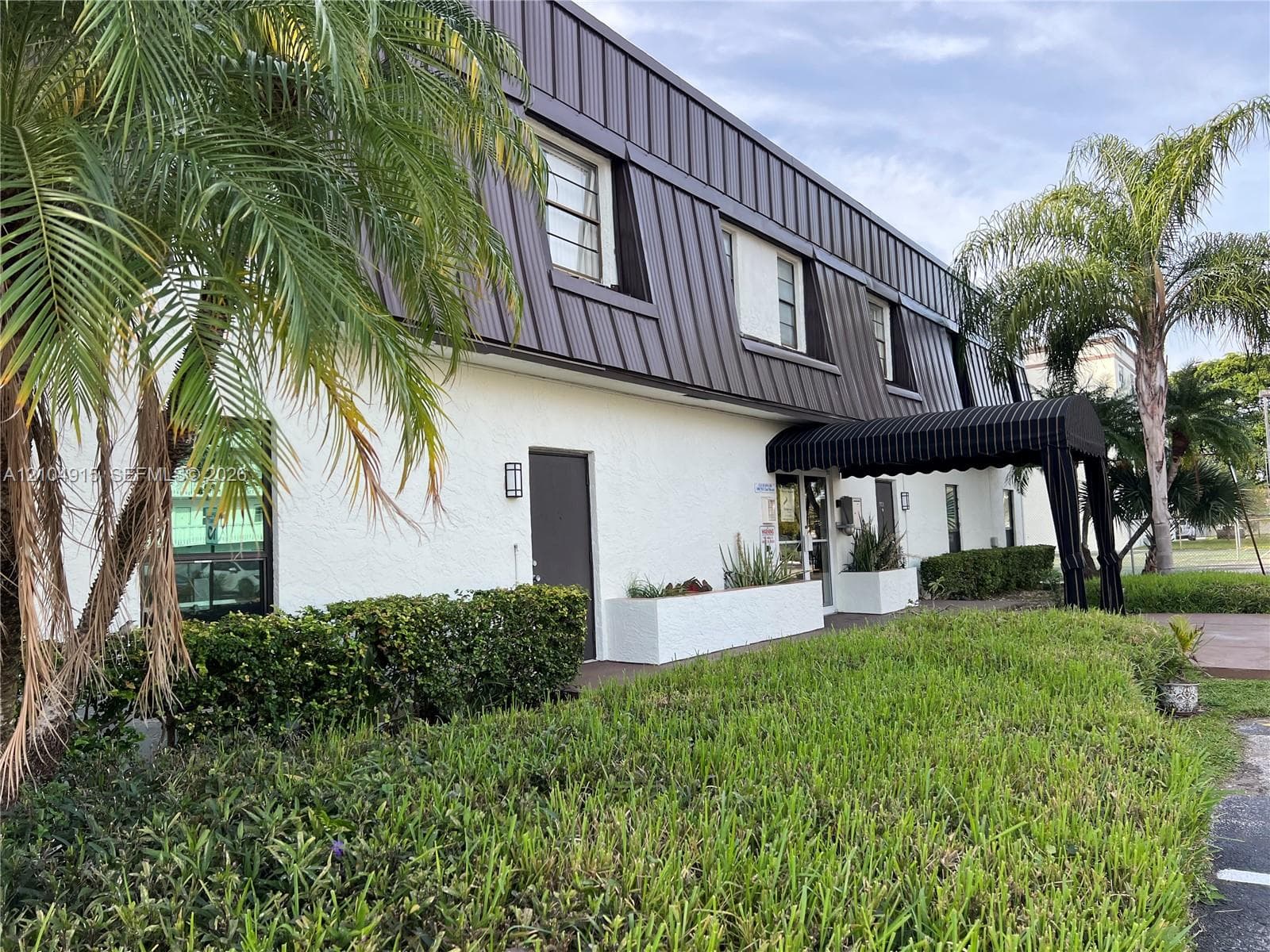Two-story exterior featuring a white stucco facade, mansard roofline with metal paneling, integrated masonry planters, and a striped fabric awning over the entry
