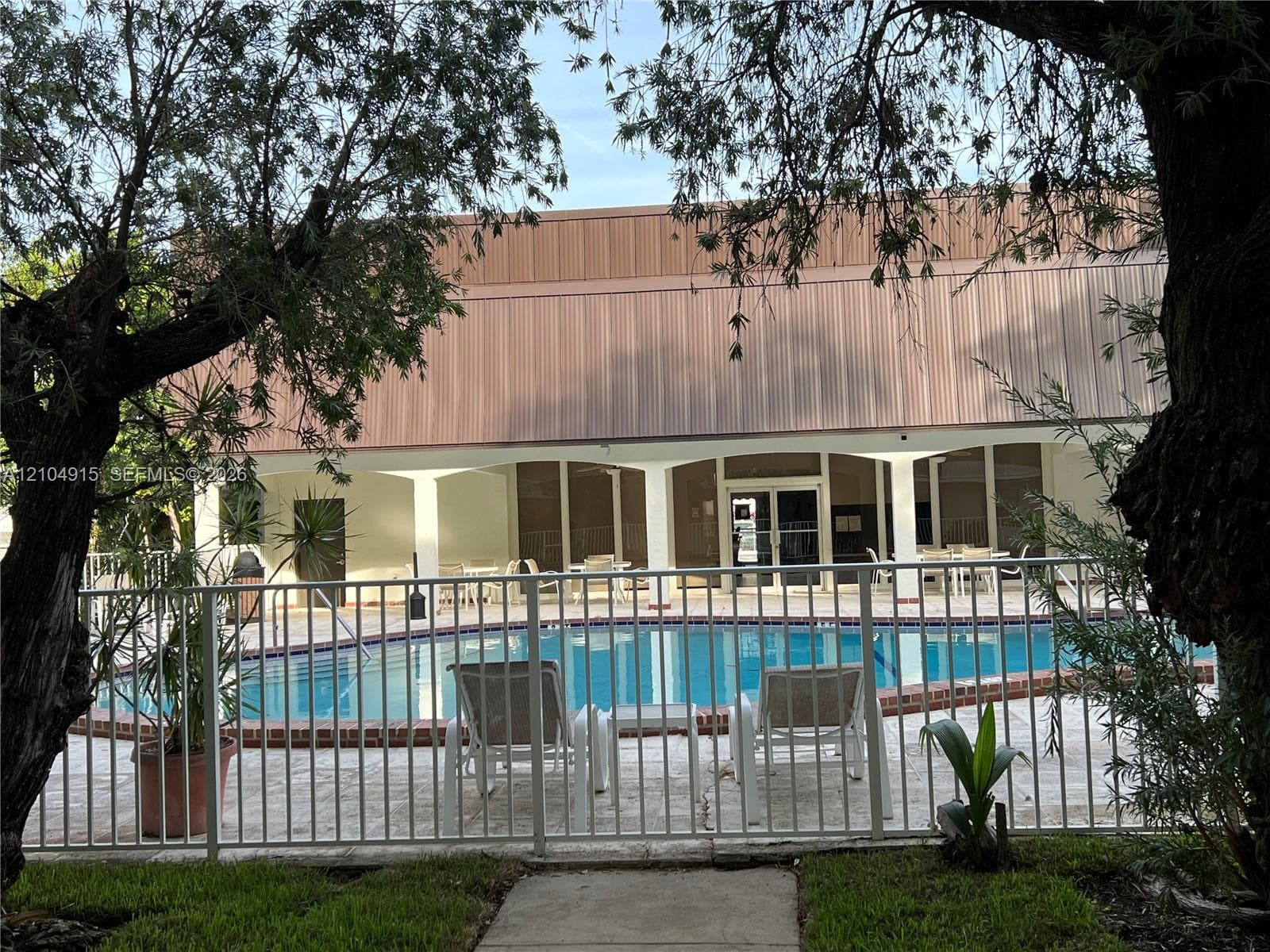 Swimming pool with brick coping, surrounded by a paved deck and enclosed by a metal perimeter fence, featuring a covered patio with structural columns
