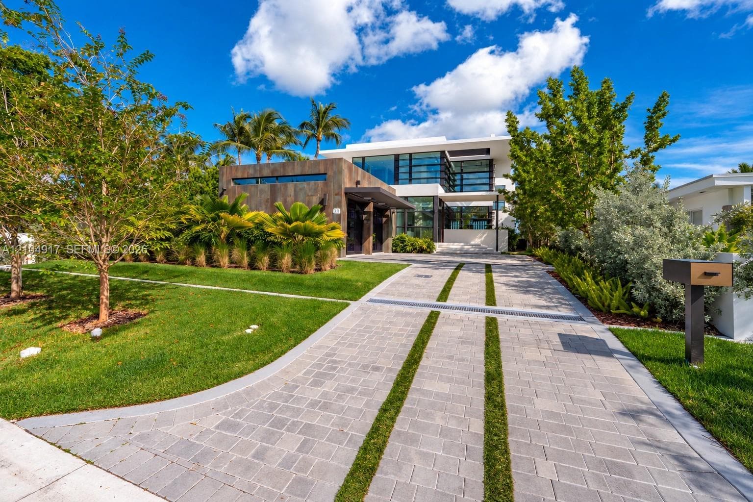 Modern architectural exterior featuring expansive glass curtain walls, horizontal stone cladding, a segmented paver driveway with integrated turf strips, and manicured landscaping