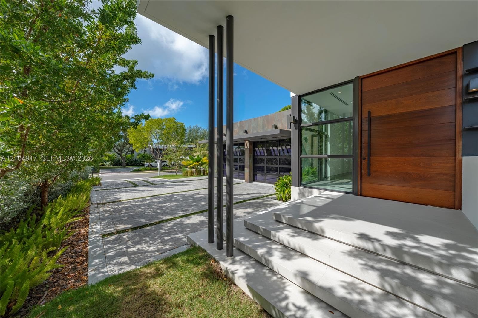 Modern entryway featuring a large wood-finish pivot door, oversized concrete steps, vertical black metal columns, and a paved driveway with grass-infill joints