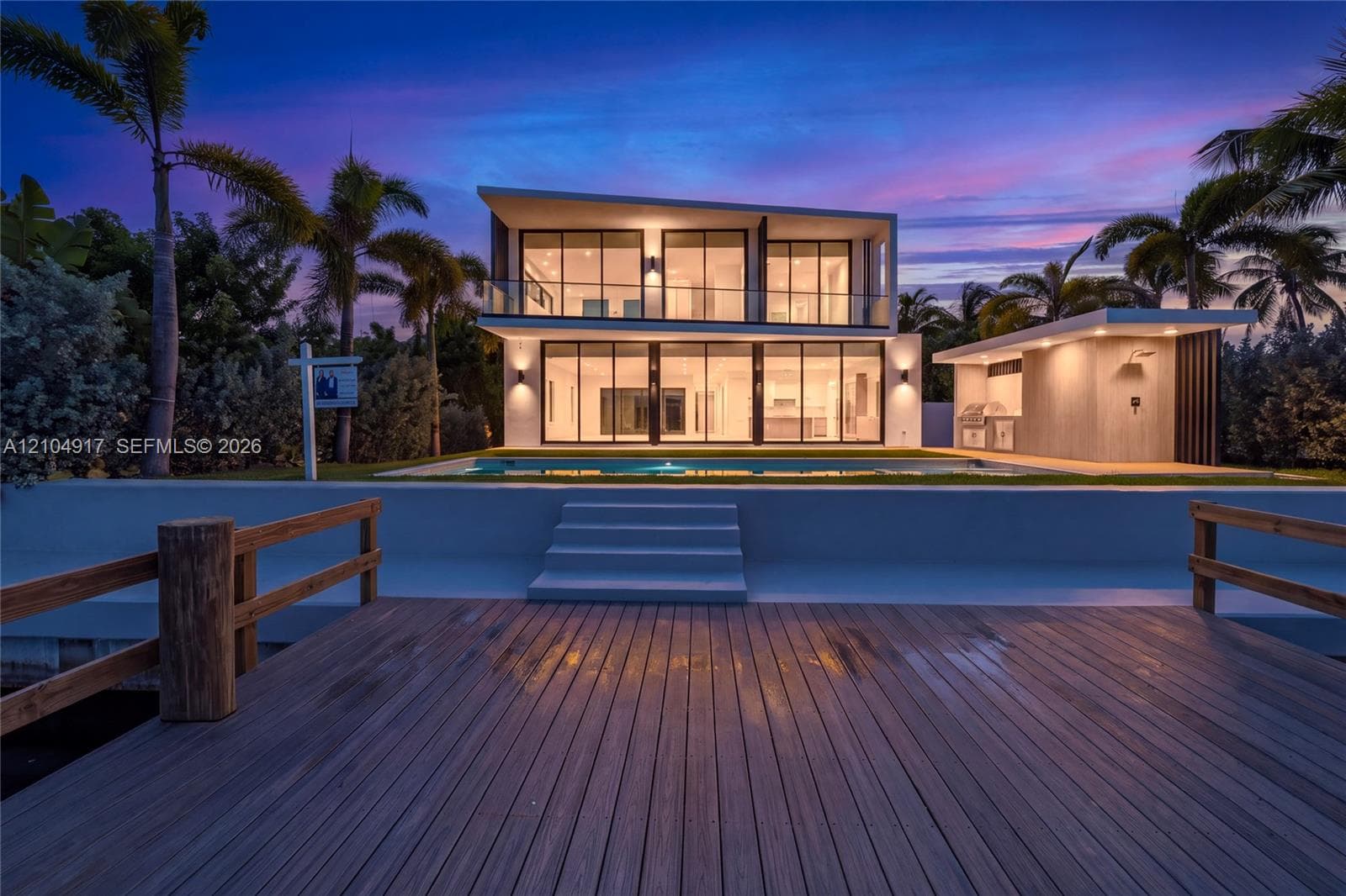 Modern two-story waterfront residence featuring floor-to-ceiling windows, a private dock with wooden decking, a built-in outdoor kitchen, and an in-ground swimming pool