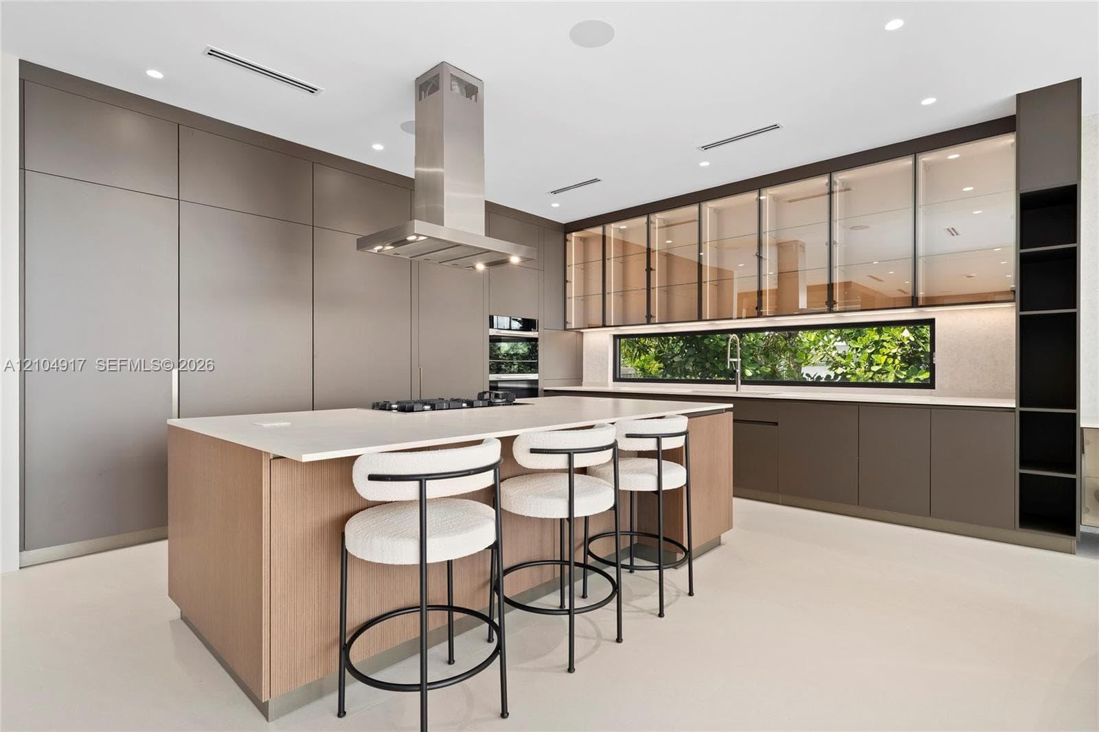 Contemporary kitchen with a large central island, stainless steel range hood, integrated appliances, and extensive cabinetry featuring glass-front uppers