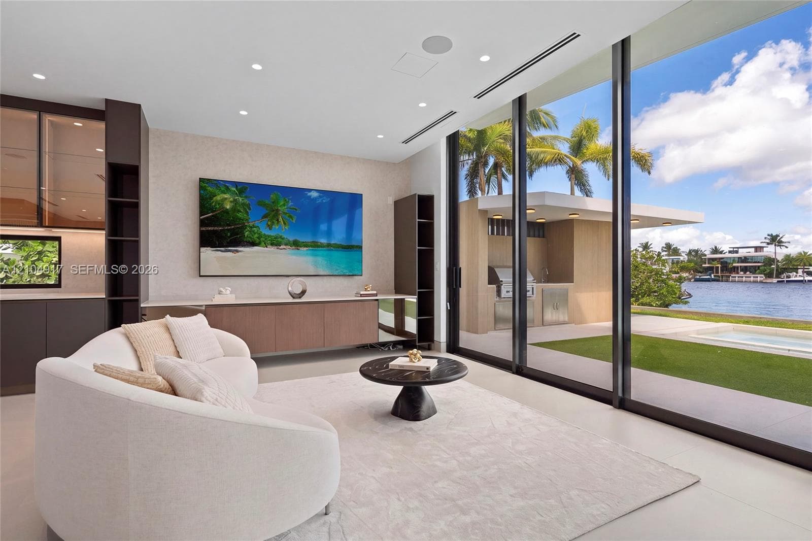 Floor-to-ceiling sliding glass doors, built-in media console, recessed lighting, integrated shelving, and an outdoor kitchen with water frontage