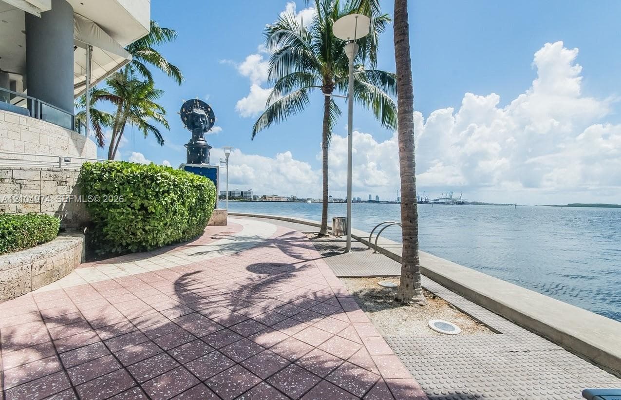 Waterfront promenade featuring patterned tile paving, stone retaining walls, manicured hedges, and mature palm trees