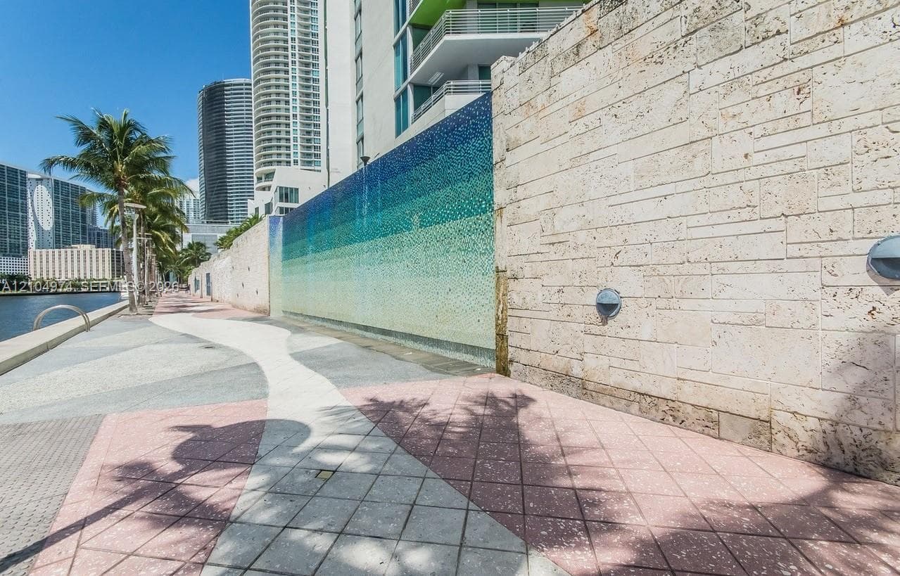 Waterfront promenade featuring a decorative mosaic tile water feature, stacked stone masonry wall, multi-toned concrete paving, and integrated exterior sconce lighting