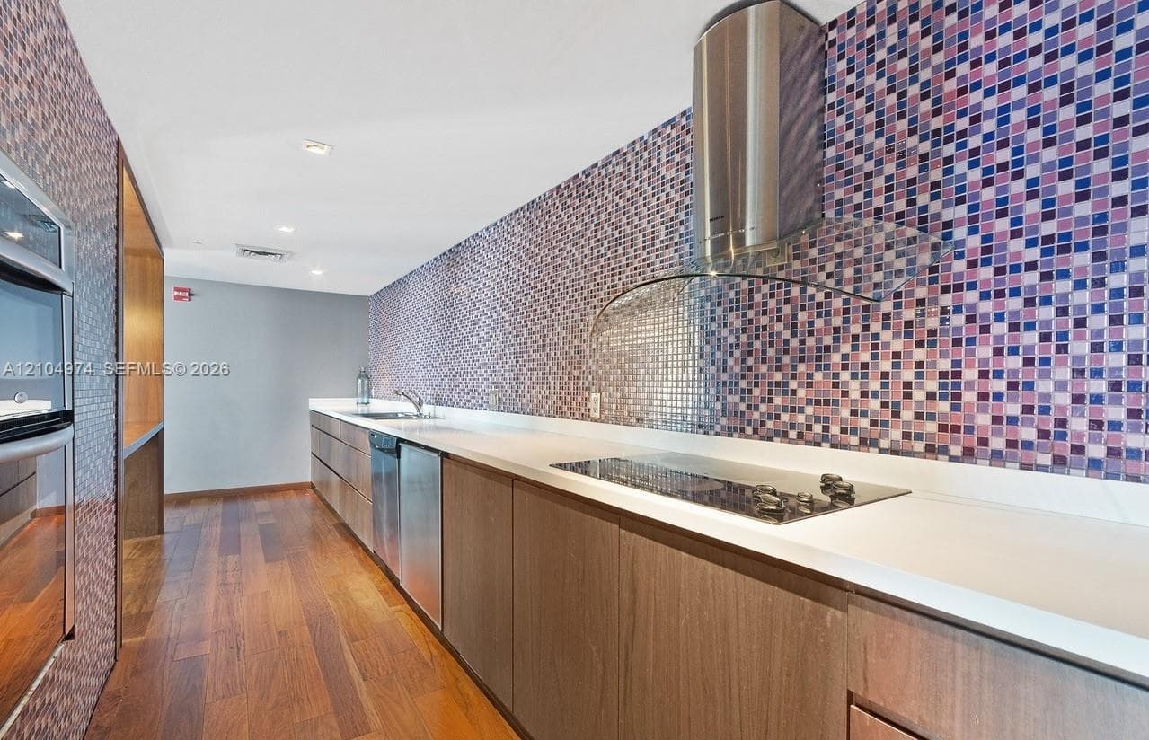 Kitchen featuring wood-finish cabinetry, white solid-surface countertops, multi-colored mosaic tile backsplash, stainless steel built-in appliances, and wood-finish flooring