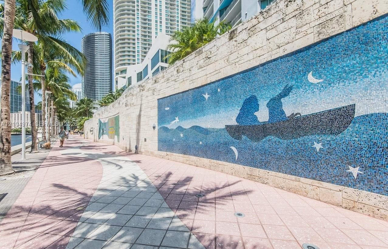 Outdoor promenade featuring decorative stone-tile wall murals, paved walkways, and palm tree landscaping