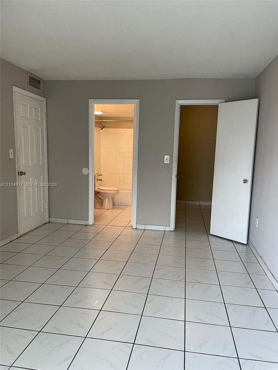 Interior room featuring white ceramic tile flooring with grey veining, three panel doors, and a bathroom containing a tiled shower enclosure and porcelain toilet