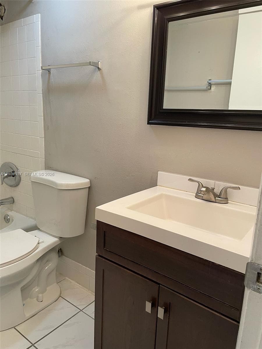 Dark wood-finish vanity with a white integrated sink, matching framed wall mirror, and tile flooring, including a white tile-surround shower combination