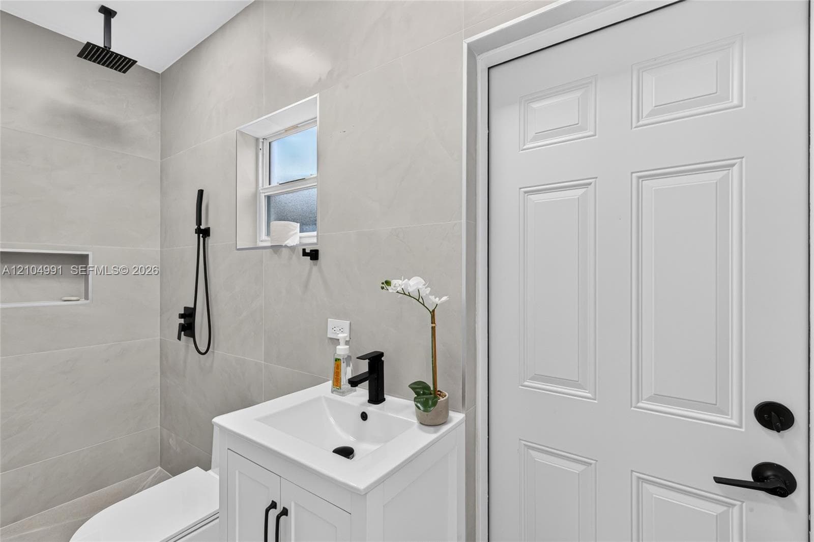 Full-height neutral wall tiling, walk-in shower with rainfall showerhead and built-in wall niche, white vanity with rectangular basin and matte black fixtures, and six-panel interior door with matte black hardware