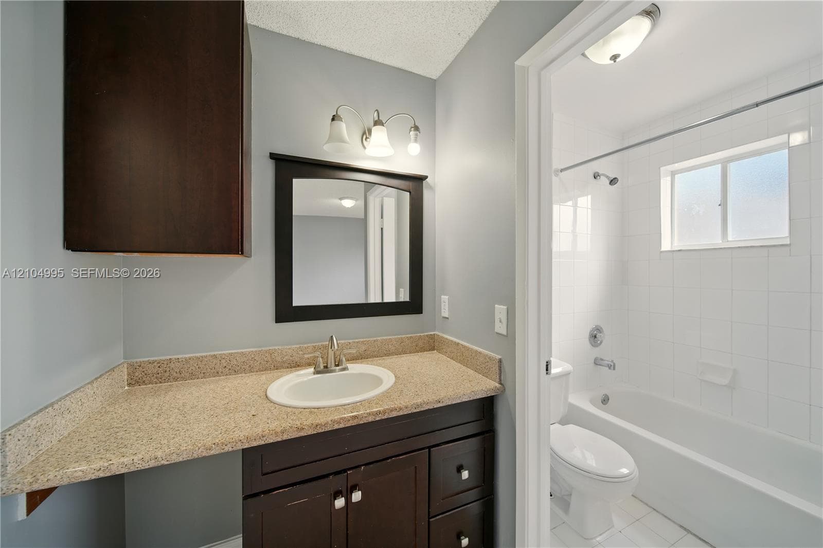 Bathroom featuring a dark-finish vanity with a stone-composite countertop, integrated sink, wall-mounted storage cabinet, and white tile shower surround