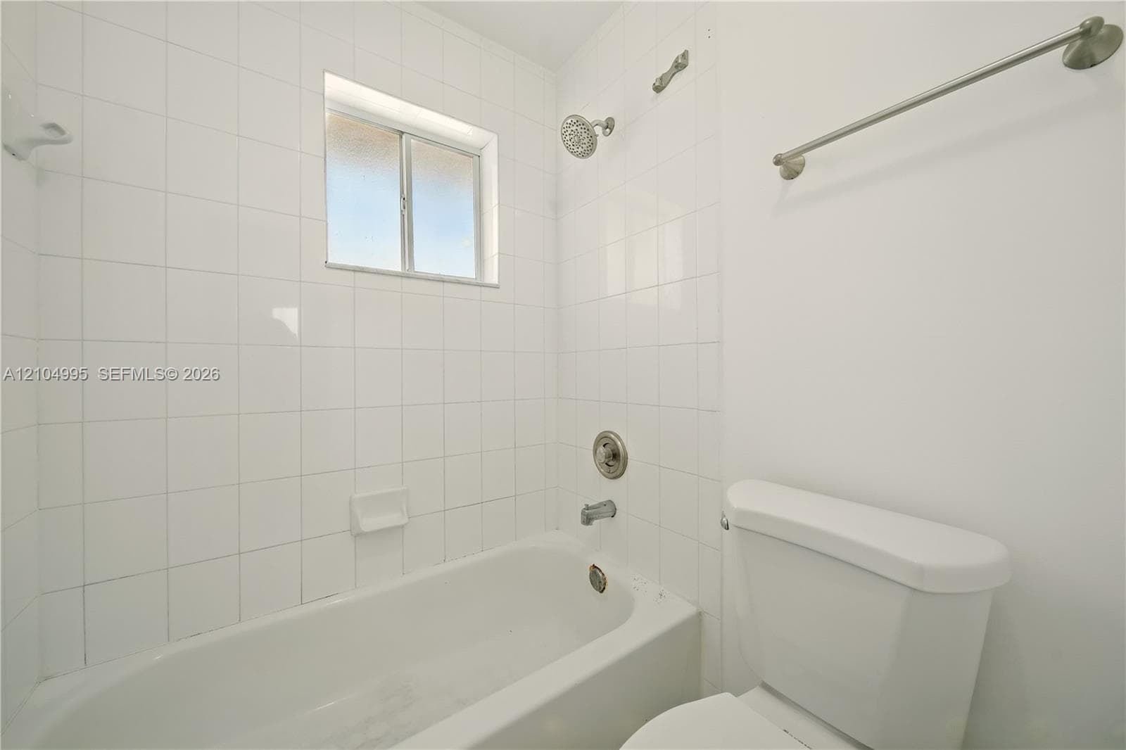Bathroom featuring full-height white wall tiling, a built-in bathtub with a brushed-nickel shower fixture, a window, a white porcelain toilet, and a wall-mounted metal towel rack