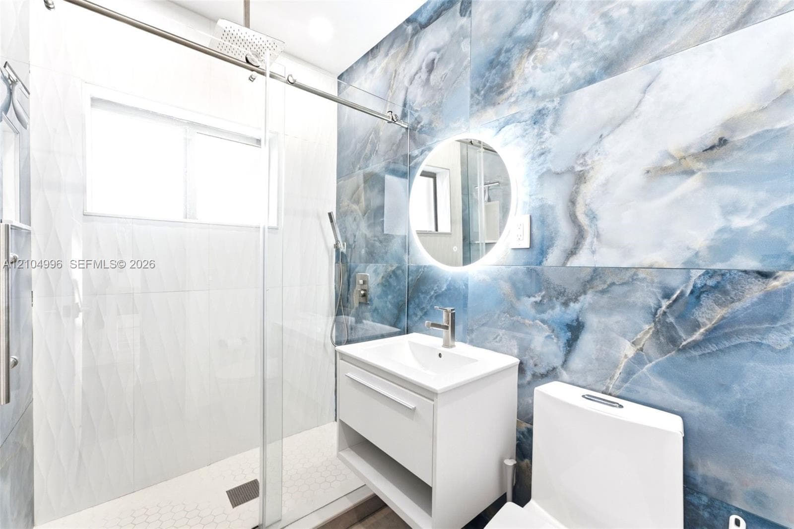 Large-format blue stone-finish wall tile, frameless glass shower enclosure with rain-style showerhead, floating vanity, circular backlit mirror, and white dual-flush toilet