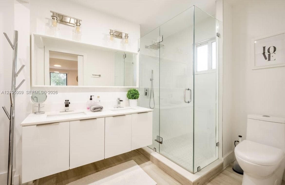 Dual sink vanity with white cabinetry and integrated sinks, large mirrored wall, glass-enclosed shower with tiled walls and floor, and wood-finish flooring