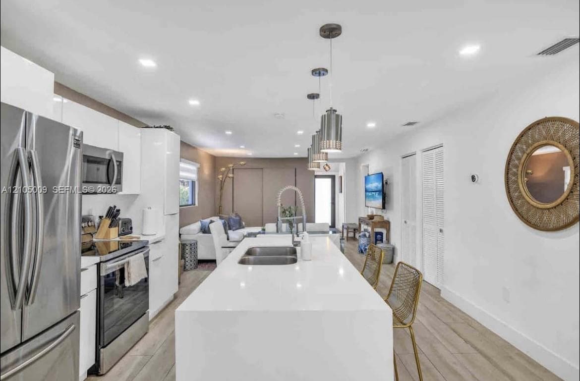Modern kitchen featuring a waterfall island with an undermount double sink, stainless steel appliances, white flat-panel cabinetry, and wood-finish flooring
